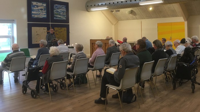 En gruppe ældre folk sidder i stoler og lytter til en foredragsholder.