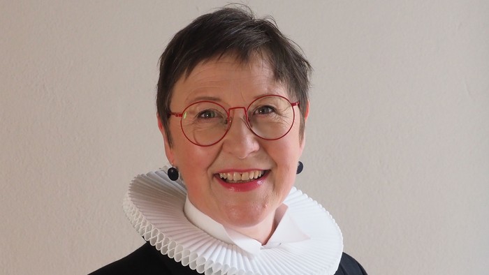 En kvinde med kort hår og røde briller, smiler og har et hvidt rødt krage under en sort jakke