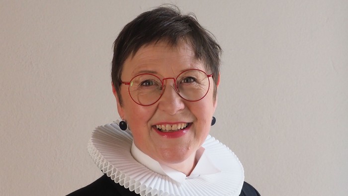 En kvinde med kort hår og røde briller, smiler og har et hvidt rødt krage under en sort jakke