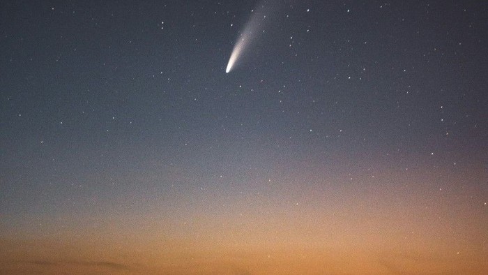 Ein Komet zieht bei Dämmerung über einem beleuchteten Ort am Himmel