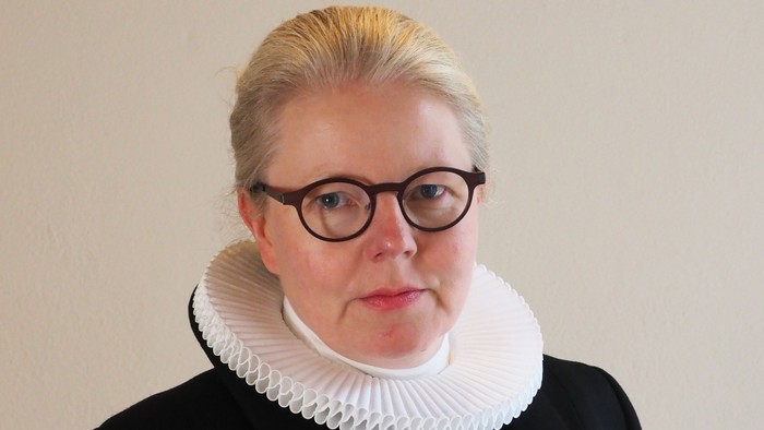 En kvinde med blond hår og briller bærer en sort jakke med hvid krage.