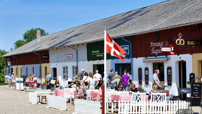 En gruppe af mennesker sidder udendørs ved bord foran et stort, traditonalt hus med en flagstang.