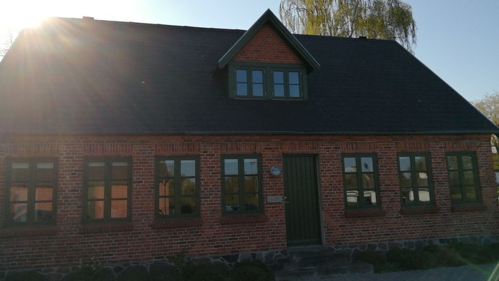 En rød murstensbygning med grøn dør og vinduer, beliggende på en stenet plads med en træ i baggrunden.