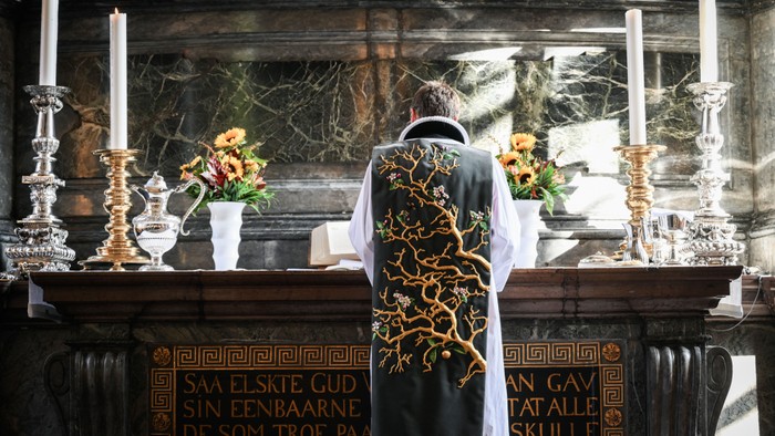 Præst i messesakkel ved alter med lysstaker og blomster.