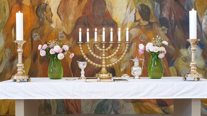 En hvid beklædning dekker bordet med en menora og grønne vaser med blomster.