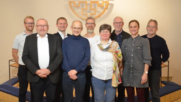 En gruppe af ni personer står sammen i en hal, alle smiler til kameraet