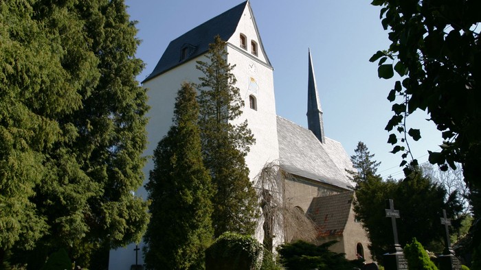 Weiße Kirche mit hohem Turm und spitzem Dach, umgeben von grünen Bäumen und Büschen