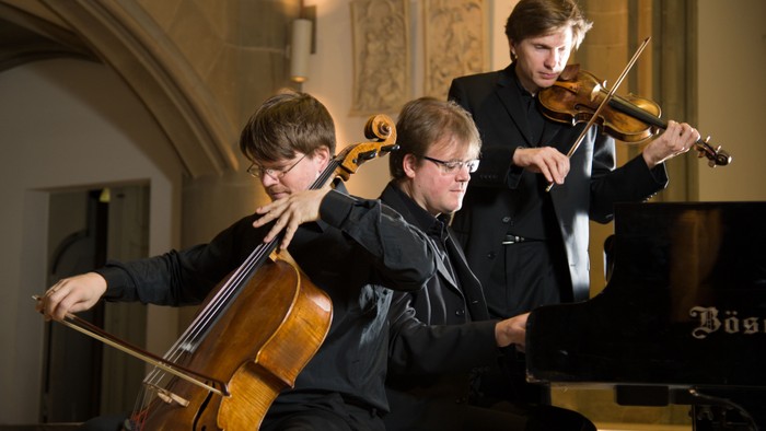 Drei Musiker spielen Cello, Klavier und Geige in einem Konzertsaal