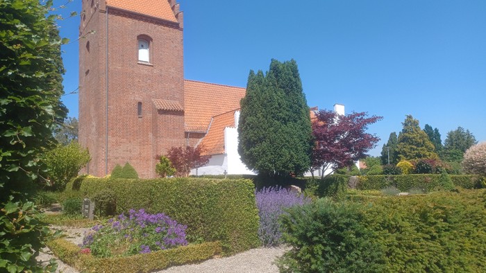 En rød murstenskirke med spiret og omgivelser med grønne buske og blomster.