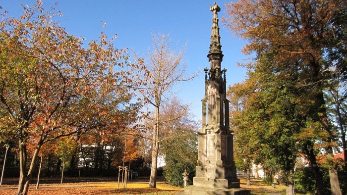 Im Park steht ein hoher, verzierter Obelisk vor Bäumen mit herbstlicher Farbgebung.