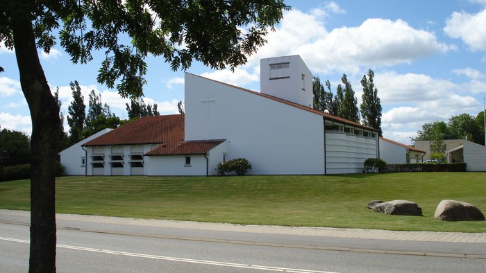 Et hvidt kirkebyggeri med rødt tag og en klokketårn, beliggende ved en vej med græs og træer i forgrunden