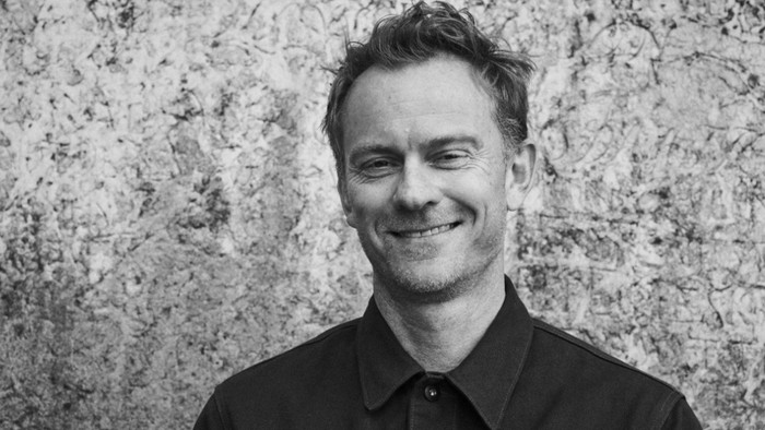 En svart-hvid portrætfoto af en mand med kort hår og skæg, der smiler og ser direkte i kameraet.