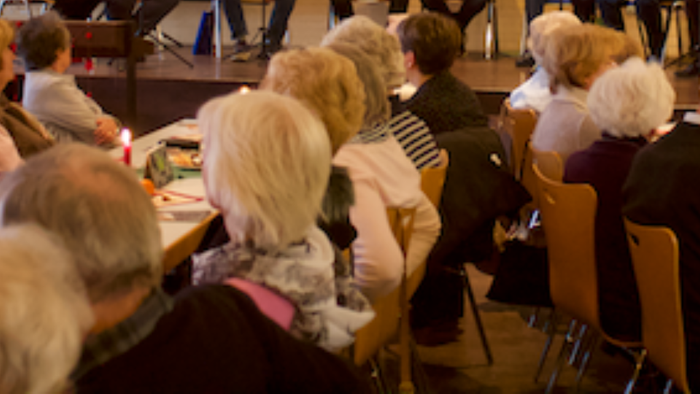 Eine Gruppe älterer Menschen sitzt in einem Raum und hört einem Vortrag zu.