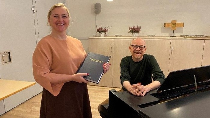 En kvinde og en mand står ved et piano, hvor kvinden holder en mappespinde.