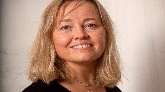 En kvinde med blondt kort hår, smilende og tørklæde bagud