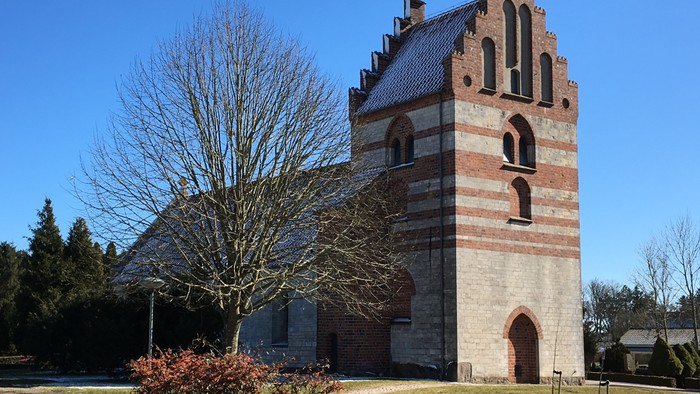 En gammel kirke med tårn og bægerhjørne vinduer, omgivet af træer og græs.