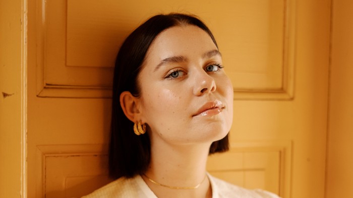 En kvinde i et hvidt krageblus og hvidt skørt står ved en gul dør.