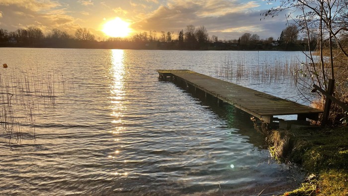 Ein Steg ragt in einen ruhigen See, die Sonne geht unter und spiegelt sich im Wasser wider