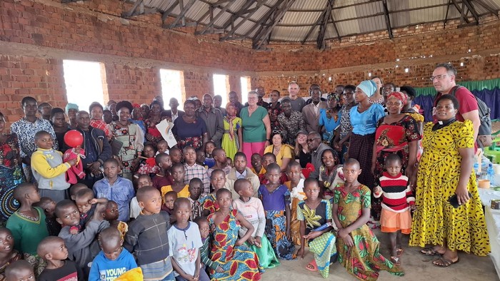Eine große Gruppe von Menschen, darunter viele Kinder, posiert in einem Raum mit Backsteinwänden und Holzboden.