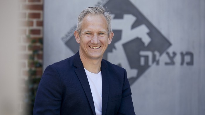 Mand i mørk blå jakke står foran mur med logo og hebraisk tekst.