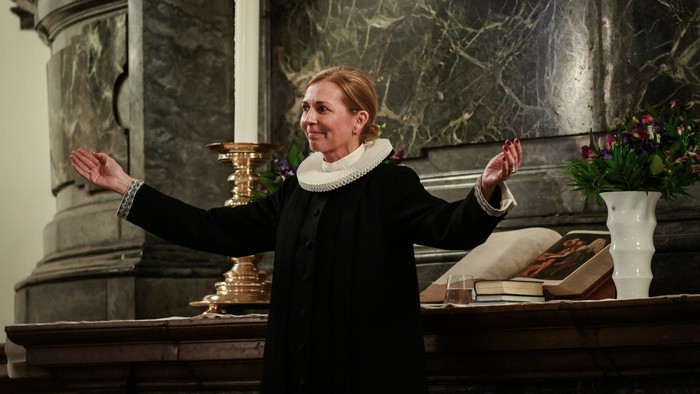 Kvinde i mørkt kapel, der holder forgyldt objekt og har arme i vejr.