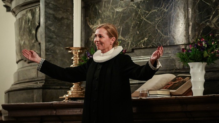 Kvinde i mørkt kapel, der holder forgyldt objekt og har arme i vejr.