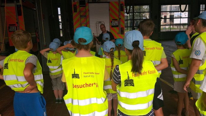 Kinder mit Warnwesten und Schutzhelmen besichtigen ein Industriegelände.