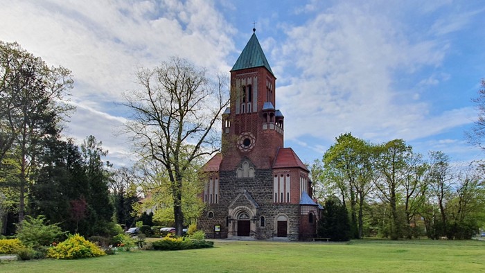 Das Bild zeigt eine rote Backsteinkirche mit hohem Turm und grünem Dach, umgeben von Bäumen und Gras.