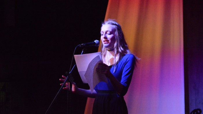 Eine Frau steht auf der Bühne und liest von einem Blatt Papier in ein Mikrofon.