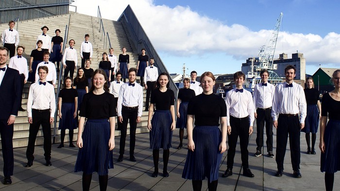 En gruppe af mænd og kvinder i formel klædning står på en terrasse med en moderne bygning i baggrunden