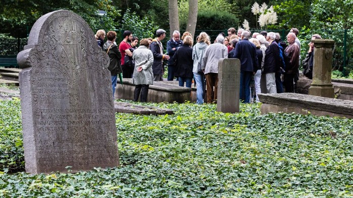 Eine Gruppe von Menschen steht in einem Friedhof und schaut sich Grabsteine an.