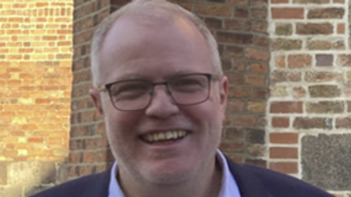 En smilende mand med briller og blå skjorte står forud for en mur af røde mursten.