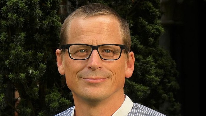 Mann mit Brille und blau-weißem Hemd vor grünem Hintergrund.