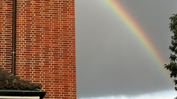 Links ein roter Ziegelbau, rechts ein Regenbogen am bewölkten Himmel