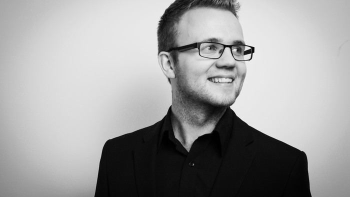 En smilende mand med briller og mørke klæder.