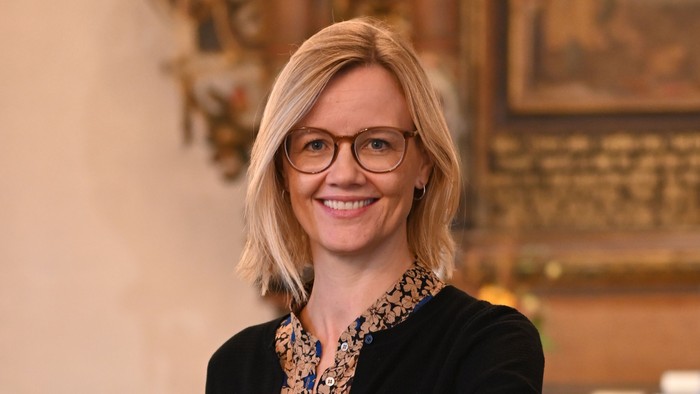 En kvinde i sort jakke og briller står ved et podium i en historisk lokalitet.