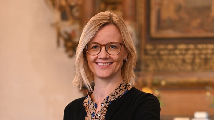 En kvinde i sort jakke og briller står ved et podium i en historisk lokalitet.