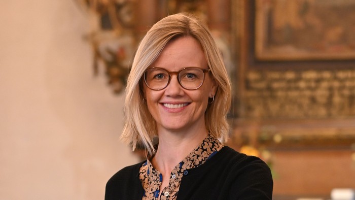 En kvinde i sort jakke og briller står ved et podium i en historisk lokalitet.