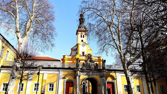 Das Bild zeigt eine gelbe Kirche mit hohem Turm und roten Türen, umgeben von kahlen Bäumen