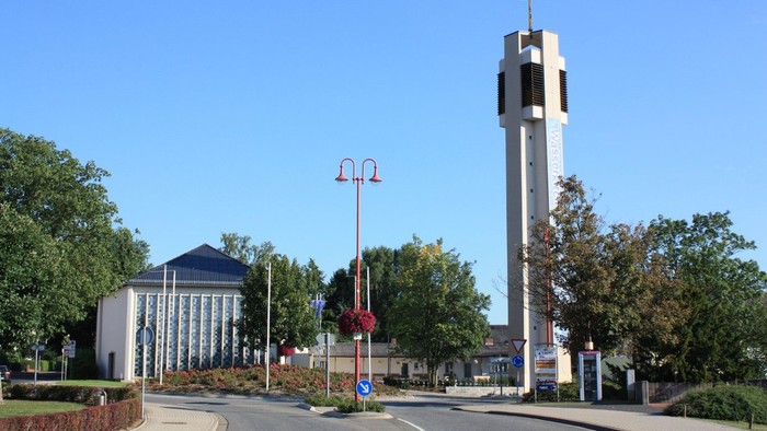 Ein hoher Kirchturm mit einem Kreuz an der Spitze steht neben einer Straße mit Bäumen und Gebäuden.
