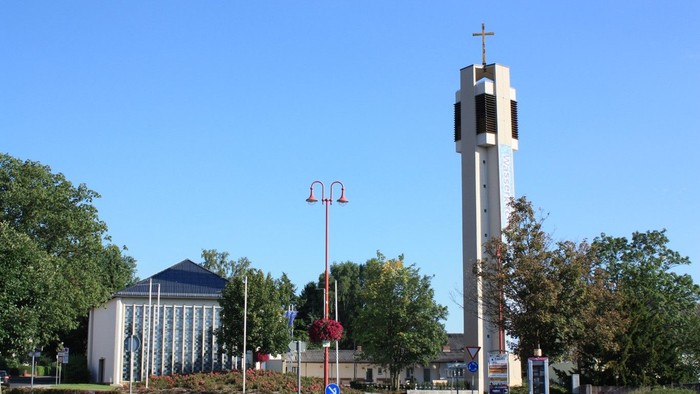 Ein hoher Kirchturm mit einem Kreuz an der Spitze steht neben einer Straße mit Bäumen und Gebäuden.