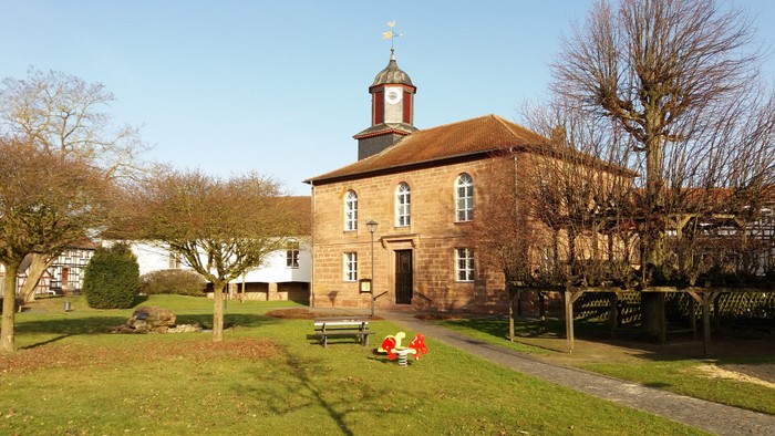 Kirche mit Kreuz auf dem Dach, Bank davor, Bäume drumherum, klarer Himmel.