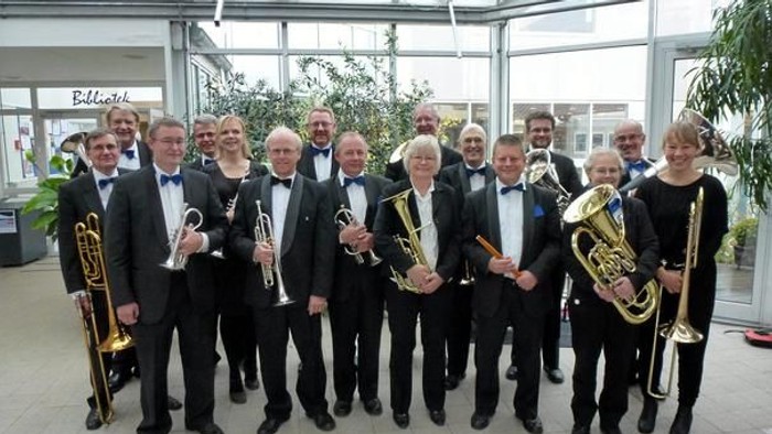 En gruppe mænd i formelle klæder holder blæseinstrumenter i en grønne omgivelser