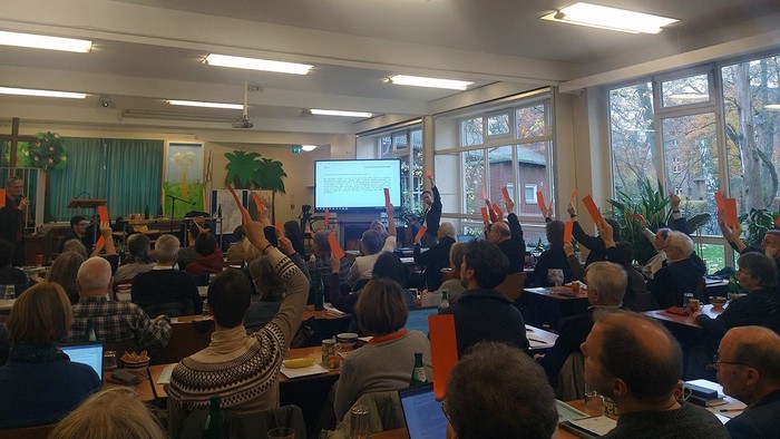 Eine Gruppe von Menschen sitzt in einem Raum und schaut auf einen Bildschirm, einige halten Orangen hoch.