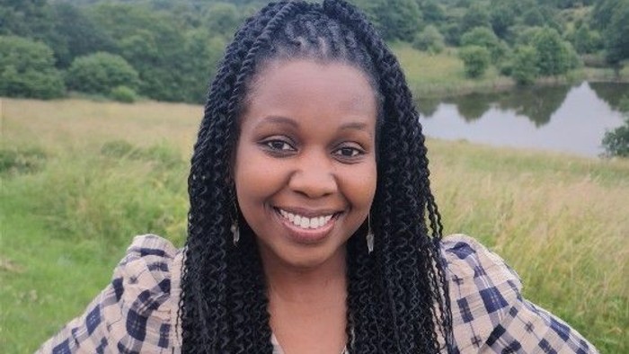 En ung kvinde med brune trætter og fløjlsjakke står i en grøn og åben naturmiljø med en sø i baggrunden.