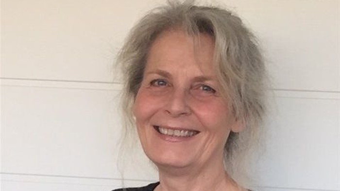 En glad kvinde med kort, grå hår står foran et hvidt, paneleret væg.