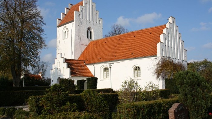 Et hvidt kirkebygning med røde tag og en tårn, omgivet af grønne buske og træer