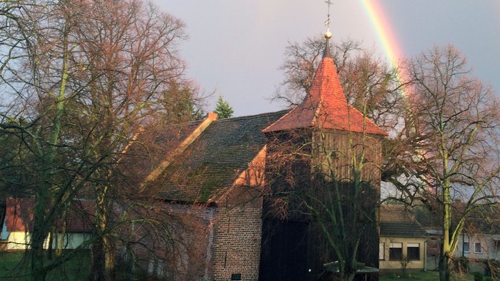 Ein Regenbogen über einem alten Backsteingebäude mit spitzem Dach und nackten Bäumen.