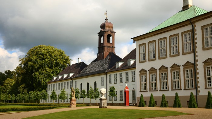 Stor hvidkalket slot med tårn og grøn tag, omgivet af græsplæne og træer