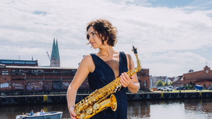 Eine Frau in einem blauen Kleid spielt Saxophon am Wasser.